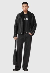 Fekete bőrkabát műszőrme gallérral, fekete grafikás póló, sötét farmernadrág, és egy fekete textil szatyor, amit az egyik kézben tart.