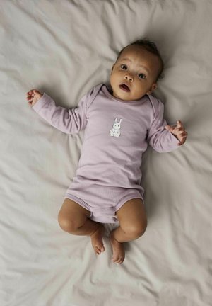 Helllila Baumwoll-Overall mit langen Ärmeln, der ein weißes Hasenmotiv auf der Vorderseite zeigt. Baby liegt auf einer beigen Stoffoberfläche.