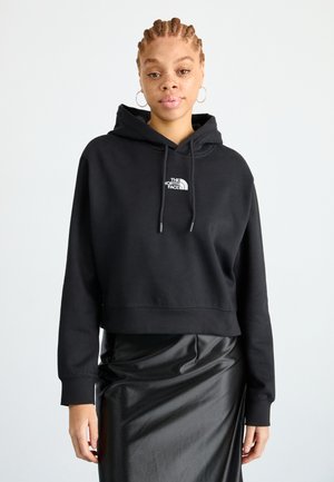 Ingekorte zwarte hoodie met een logo aan de voorkant. Voorzien van een capuchon met trekkoord en lange mouwen. Gecombineerd met een strakke, glanzende zwarte rok.