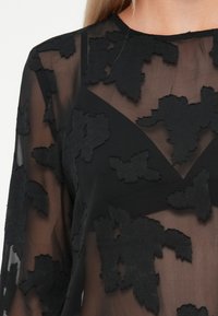 Sort gennemsigtig top med teksturerede blomsterapplikationer, lange ærmer og rund halsudskæring, der fremhæver et lagdelt og dimensionalt design.