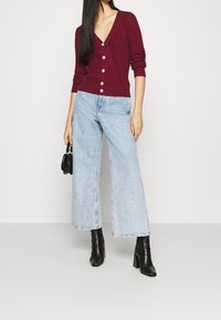 Žena nosící burgundy zapínací cardigan, světle modré široké džíny, černé boty na podpatku, držící malou černou kabelku.