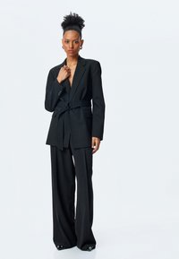 Blazer negro ajustado con solapas anchas y cintura ceñida, combinado con pantalones negros de talle alto y corte holgado. Tejido suave, con detalles mínimos.