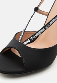 Μαύρο πέδιλο με ψηλό τακούνι, ανοιχτά δάχτυλα, μπεζ πάτο και μαύρα λουράκια με λευκή επιγραφή "LOVE MOSCHINO".