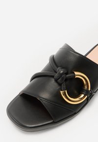Svarte lær slide sandaler med knyttet remdesign og en gull sirkulær detalj. Sålen er flat med firkantet tå.