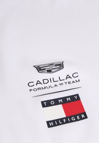 Το λογότυπο της ομάδας Formula 1 της Cadillac πάνω από μια ορθογώνια σημαία Tommy Hilfiger με κόκκινους, λευκούς και ναυτικού μπλε τόνους σε λευκό φόντο.