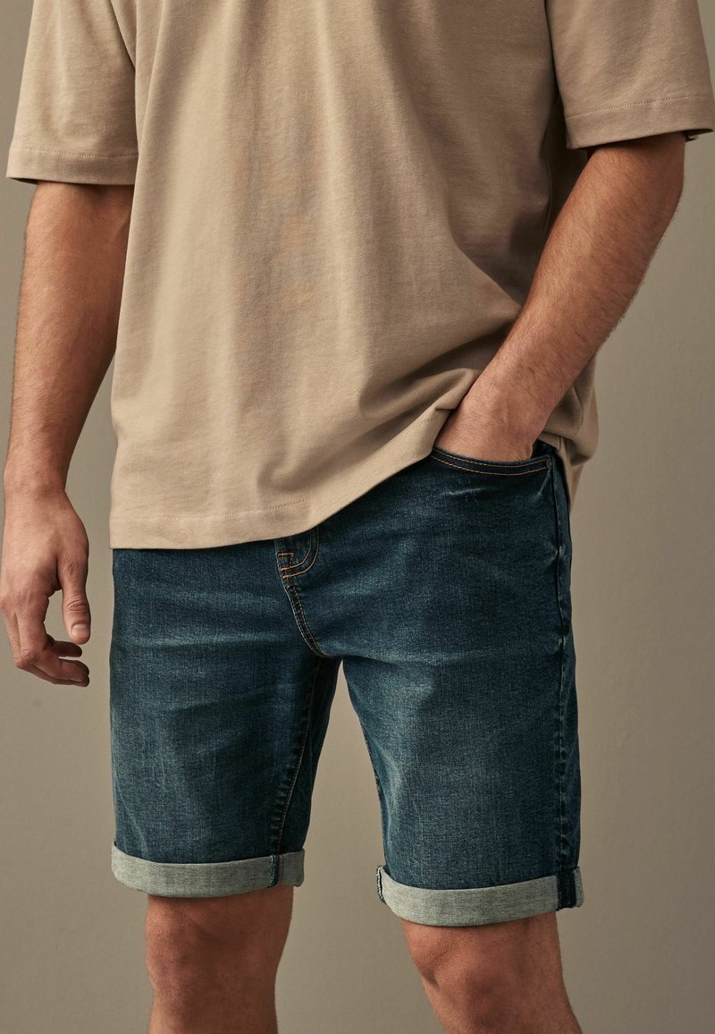 Donkerblauwe denimshorts met opgerolde mouwen, gecombineerd met een lichtbeige shirt met korte mouwen. De stof lijkt zacht en heeft een casual pasvorm.