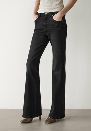 Persona con jeans neri a vita alta e gamba larga, mani in tasca, e tacchi marroni con punta quadrata su un pavimento liscio.