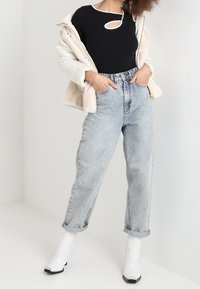 Top negro con detalle recortado, chaqueta de piel sintética color crema, jeans de tiro alto en azul claro y botas blancas hasta el tobillo con punta cuadrada.