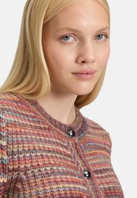Blonde woman with blue eyes wearing a multicolored striped knit sweater with large black buttons on a white background.