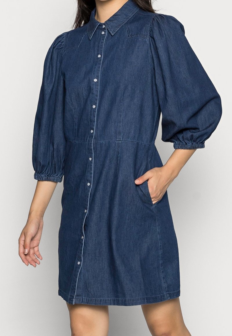 Denim dress with a collared neckline, puffed short sleeves, button front, and side pockets. Features a dark blue color and soft texture.