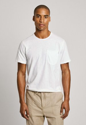 T-shirt bianca in cotone con maniche corte e taschino sul lato sinistro, abbinata a pantaloni beige, caratterizzati da una vestibilità rilassata e un design semplice.