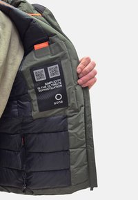 Interno di un giubbotto imbottito verde con rivestimento nero, include un'etichetta "SAFE POCKET", codici QR e una toppa di testo che recita "La semplicità è la massima sofisticazione."