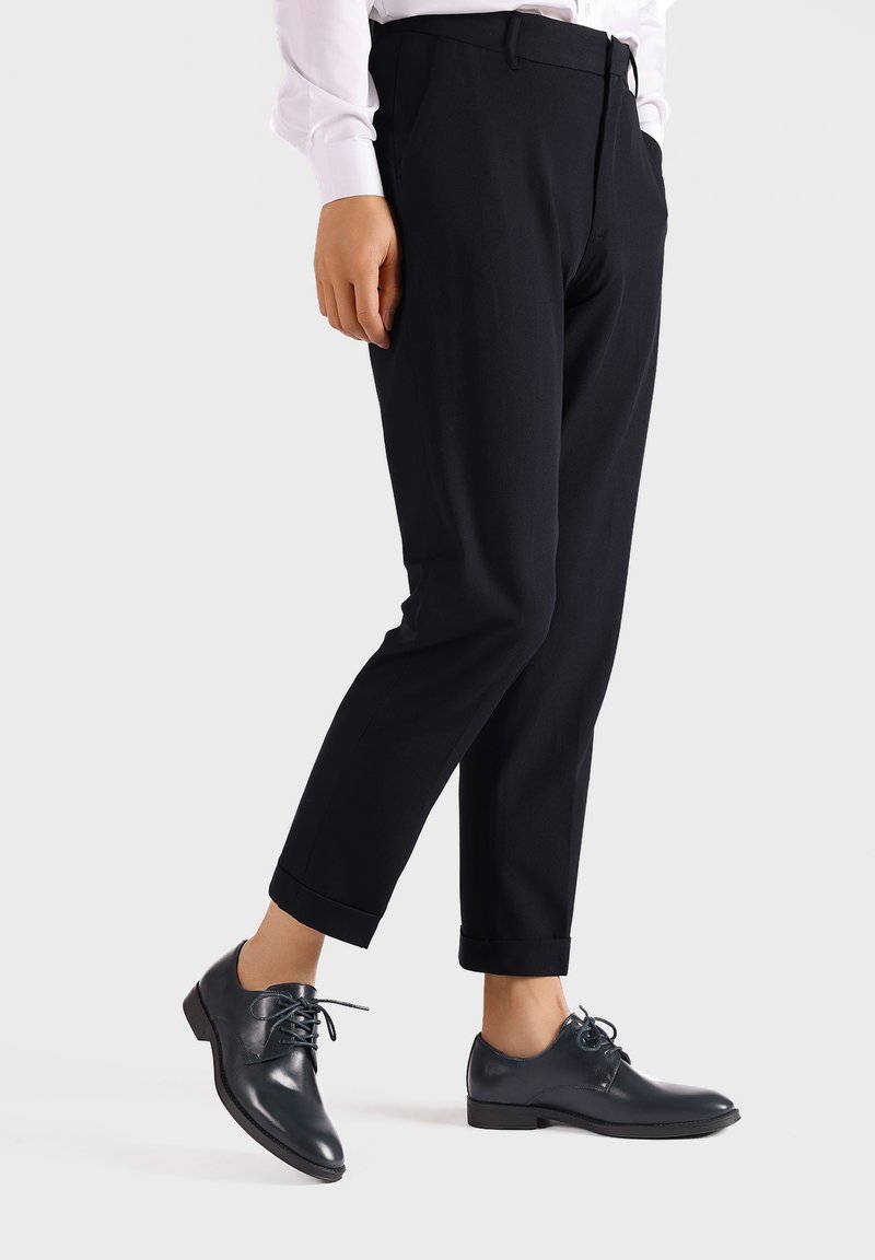 Pantalones negros ajustados con un corte slim y puños enrollados, combinados con zapatos de cuero oscuro con cordones. Tela suave, diseño minimalista.