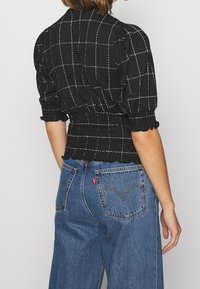 Sort struktureret bluse med hvidt rutenetmønster, puffærmer og elastisk talje, parret med højtaljede blå denimjeans.