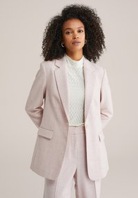 Šviesiai rožinis pritaikytas švarkas su įpjautomis atlapomis, priekinėmis kišenėmis ir vienu sagos užsegimu. Dėvimas ant tekstūruoto balto topelio.