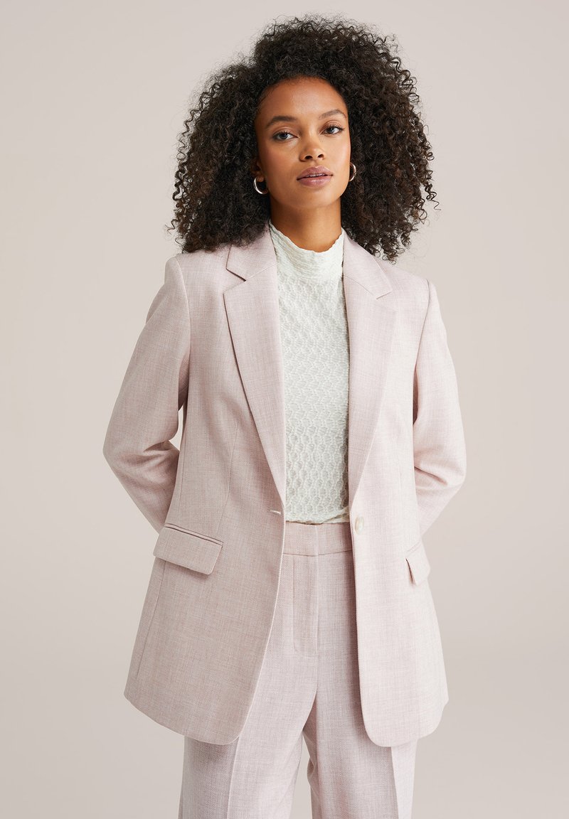 Šviesiai rožinis pritaikytas švarkas su įpjautomis atlapomis, priekinėmis kišenėmis ir vienu sagos užsegimu. Dėvimas ant tekstūruoto balto topelio.