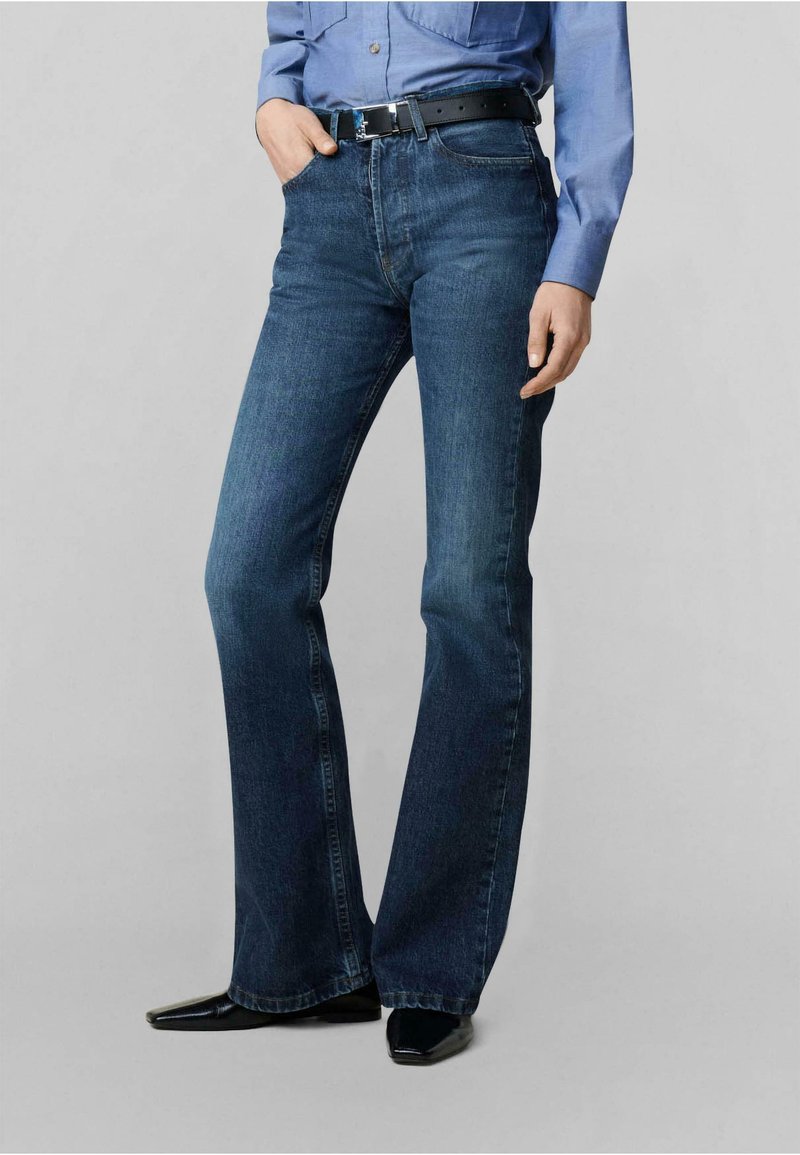 Dunkelblaue Denim-Jeans mit Schlag, eng an der Taille, vorderen Taschen und sichtbaren Nähten. Kombiniert mit einem schwarzen Leder-Gürtel und spitzen Schuhen.