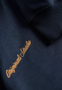 Μπλέ σκούρο φούτερ με ριγωτές μανσέτες; διαθέτει χρυσό κεντημένο κείμενο που γράφει "Originals Studio" μπροστά.