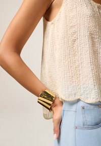 Brazalete de oro con una superficie pulida y reflectante, llevado sobre una blusa de crema clara texturizada con un dobladillo con escote, combinado con jeans azul claro.