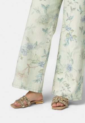 Picioare purtând pantaloni largi cu model floral deschis și sandale bej cu vârf deschis, decorate cu elemente aurii și cristale, pe un fundal alb.