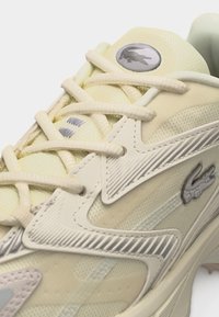 Ανοιχτό μπεζ αθλητικό παπούτσι με κρέμ κορδόνια, υφασμένα επικάλυψη και ασημένια λογότυπα κροκόδειλου στη γλώσσα και την πλευρική επιφάνεια.