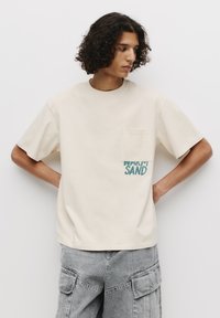 Beige bomulds t-shirt med rund hals, der har en lomme og blå tekst "WARM SAND" på forsiden. Modellen har grå cargo bukser på.