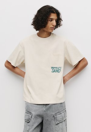 Beige bomulds t-shirt med rund hals, der har en lomme og blå tekst "WARM SAND" på forsiden. Modellen har grå cargo bukser på.