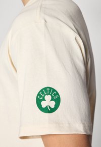 Krem bomull t-skjorte med en grønn, sirkulær logo som har "CELTICS" og et design av en trebladet kløver på venstre erme.