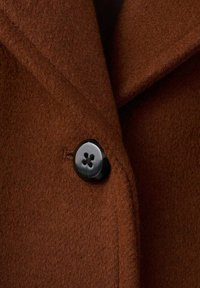 Bruine wollen jas met een zachte textuur, voorzien van een zwarte knoop met vier gaatjes en een reverskraag. Zichtbare stikseldetails.