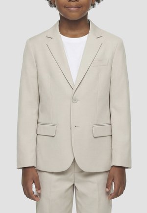 Bambino che indossa una giacca da abito beige e pantaloni abbinati sopra una camicia bianca, in piedi davanti a uno sfondo grigio chiaro uniforme.