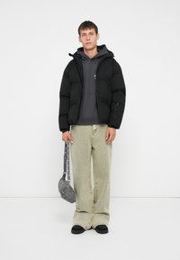 Svart dunjakke med hette, grå sweatshirt under, lysegrønne vide bukser og en liten grå veske med stropp.