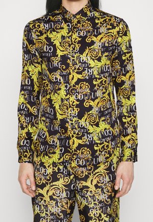 Sort skjorte med knapper og bukser med gule barokblade-mønstre og hvid "Versace Couture"-tekst båret af person med langt sort hår.