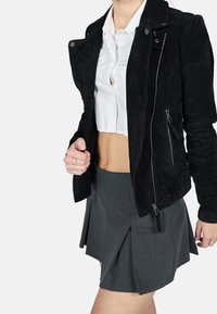 Černá semišová bunda se zipy a patentkami, nošená přes zkrácenou bílou košili s knoflíky, v kombinaci se šedou plisovanou sukní.