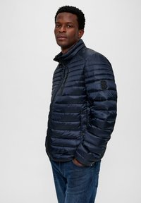 Marineblauwe pufferjack met verticale gestikte patroon, hoge kraag en ritssluiting. Heeft een zwart logoaccent op de mouw. G gedragen met jeans.