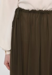 Olíva zöld, pliszírozott szoknya könnyű anyagból, rugalmas derékkal és zsinóros zárással. Simaság jellemzi, áramló dizejn.