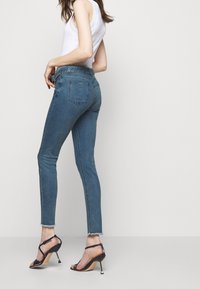 Smal blå denimjeans med fransad fåll, med bakfickor, bärs tillsammans med svarta högklackade sandaler. Toppen är en vit linne.