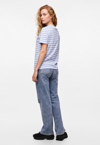 Striped t-skjorte i lyseblå og hvit bomull, kombinert med lyseblå jeans og svarte kraftige sko. Modellen vises fra baksiden.
