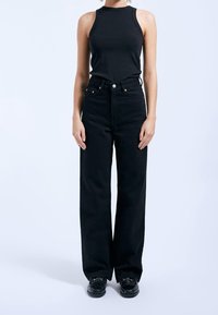 Svart ärmlös topp kombinerad med svart, högmidjad, löst sittande jeans. Särskilda detaljer inkluderar kopparaccents och knappstängning.