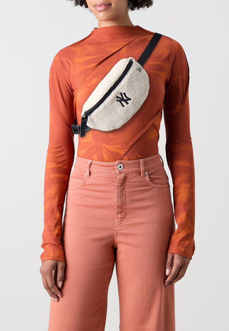 Vit fleece crossbody-väska med svart dragkedja och logotyp, bärs över en orange långärmad topp och rosa byxor, som visar upp en avslappnad stil.