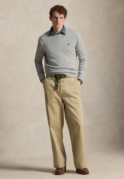 Man draagt een grijze trui over een blauw overhemd met kraag, beige broek met een groene riem met patroon en bruine schoenen, staat met handen in de zakken.