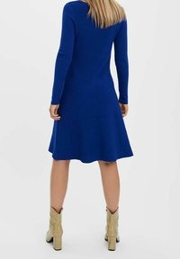 Robe bleue à manches longues avec un ourlet évasé, texture tricotée et col rond. Portée avec des bottines beiges texturées. Vue de dos.