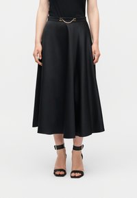 Γυναίκα που φοράει μαύρη μέση φούστα με λεπτή ζώνη με χρυσές λεπτομέρειες, συνδυασμένη με μαύρες γόβες με λουράκι στον αστράγαλο και ανοιχτά δάχτυλα, σε λευκό φόντο.