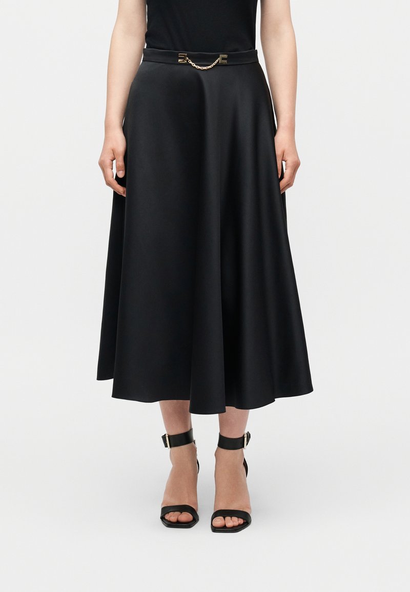 Γυναίκα που φοράει μαύρη μέση φούστα με λεπτή ζώνη με χρυσές λεπτομέρειες, συνδυασμένη με μαύρες γόβες με λουράκι στον αστράγαλο και ανοιχτά δάχτυλα, σε λευκό φόντο.