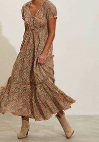 Hnědé maxi šaty s krátkými rukávy a zúženým pasem, doplněné béžovými kotníkovými botami. Tkanina má složité vzory.