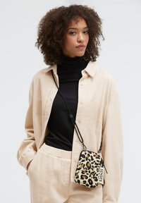 Umhängetasche mit Leopardenmuster, Reißverschluss, verstellbarem Riemen und Textetikett. Das Modell trägt ein strukturiertes beiges Hemd und darunter einen schwarzen Rollkragenpullover.