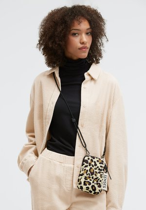 Umhängetasche mit Leopardenmuster, Reißverschluss, verstellbarem Riemen und Textetikett. Das Modell trägt ein strukturiertes beiges Hemd und darunter einen schwarzen Rollkragenpullover.