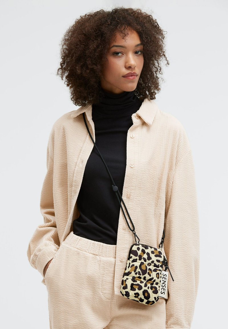 Umhängetasche mit Leopardenmuster, Reißverschluss, verstellbarem Riemen und Textetikett. Das Modell trägt ein strukturiertes beiges Hemd und darunter einen schwarzen Rollkragenpullover.