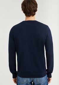 Man med brunt hår som bär en marinblå långärmad tröja och blå jeans, sedd bakifrån mot en enkel vit bakgrund.