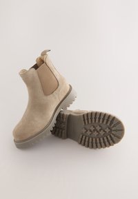 Beige ruskind ankelstøvler med elastiske sidepaneler, en trækskive og teksturerede gummisåler med en slidbane for bedre greb.