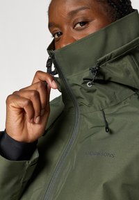Veste imperméable verte avec un col montant, une fermeture éclair à l'avant et des cordons de serrage ajustables. Présente un logo près de la fermeture éclair et des poignets noirs.